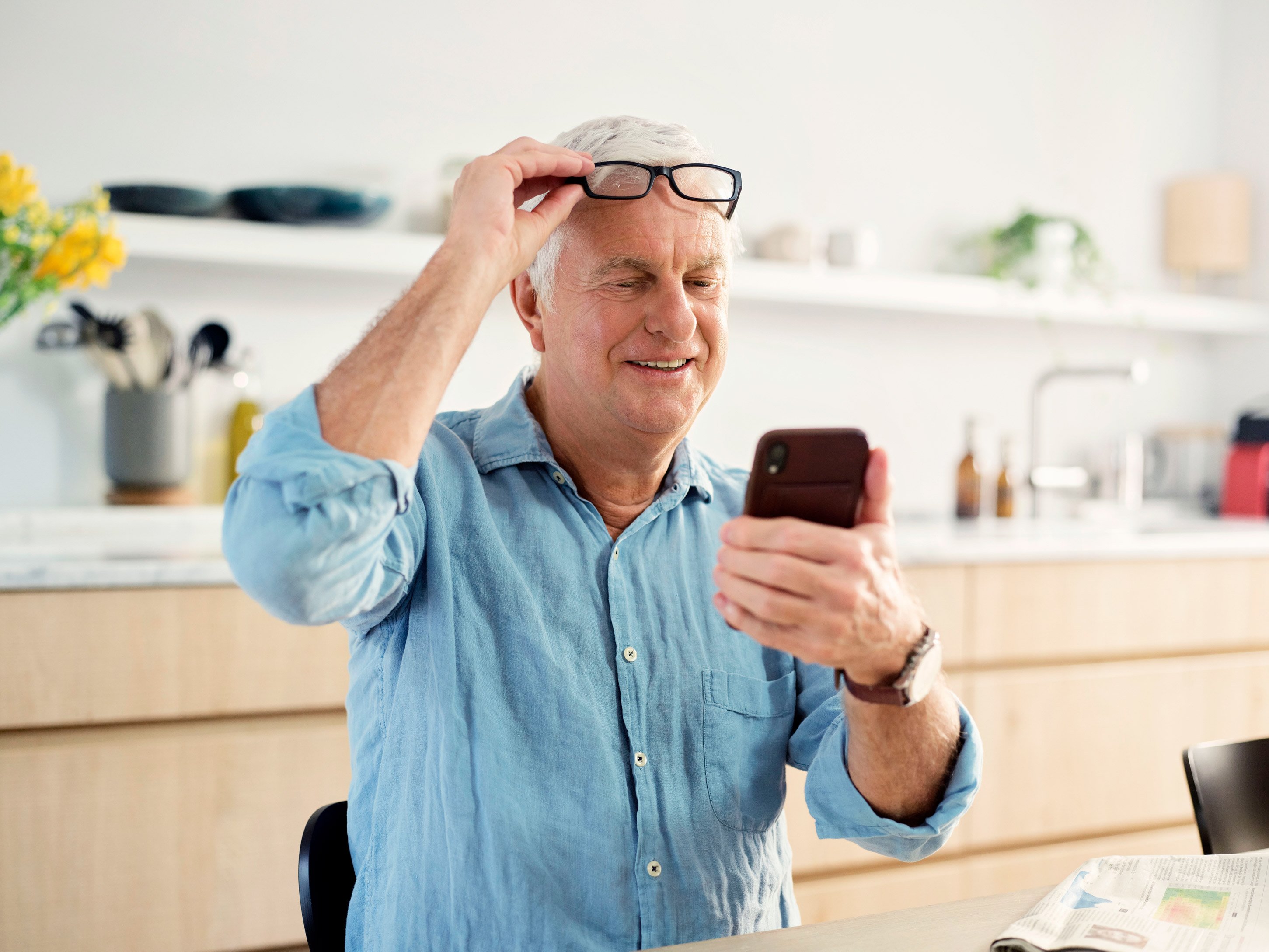 An elderly gentleman holding a phone in his left hand and looking at it while adjusting his glasses with his right hand